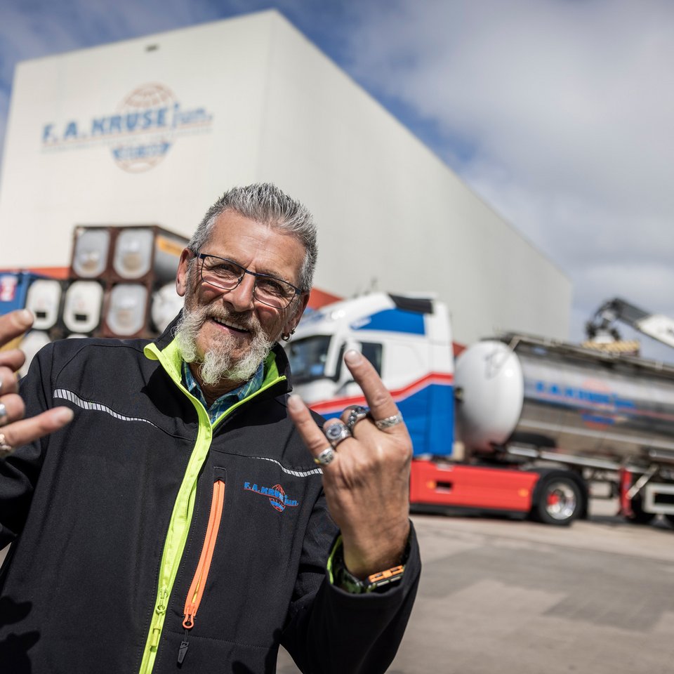 Mann mit Sonnenbrille zeigt Handzeichen vor einer großen Lagerhalle und einem Tanklastzug.