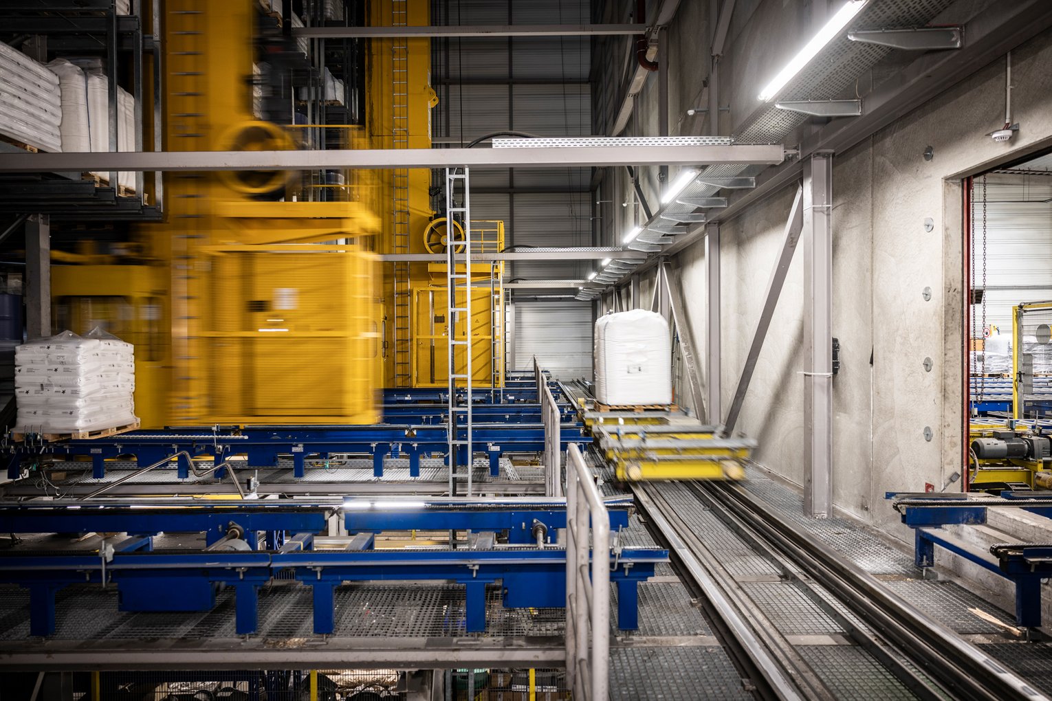 Automatisiertes Fördersystem mit gelbem Regalbediengerät und Paletten in einem Hochregallager.