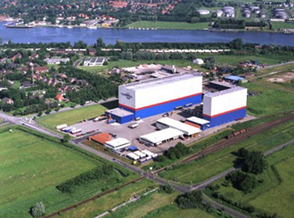 Luftaufnahme eines modernen Industriekomplexes mit Hochregallager, Bahngleis und Fluss im Hintergrund.