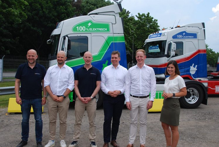 Sechs Personen stehen vor zwei bunten E-LKW mit grün-blauem und blau-rotem Design.