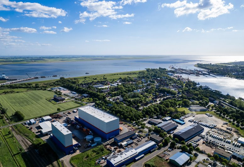 Luftaufnahme eines Logistik- und Industriegebiets mit Hochregallagern, Feldern und Flusslandschaft bei Sonnenschein.