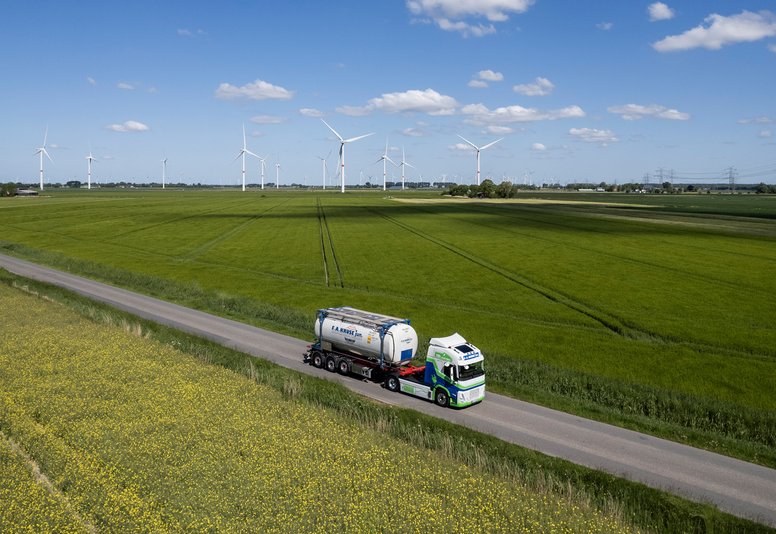 Tanklastzug fährt auf einer schmalen Landstraße durch grüne Felder mit Windrädern im Hintergrund.