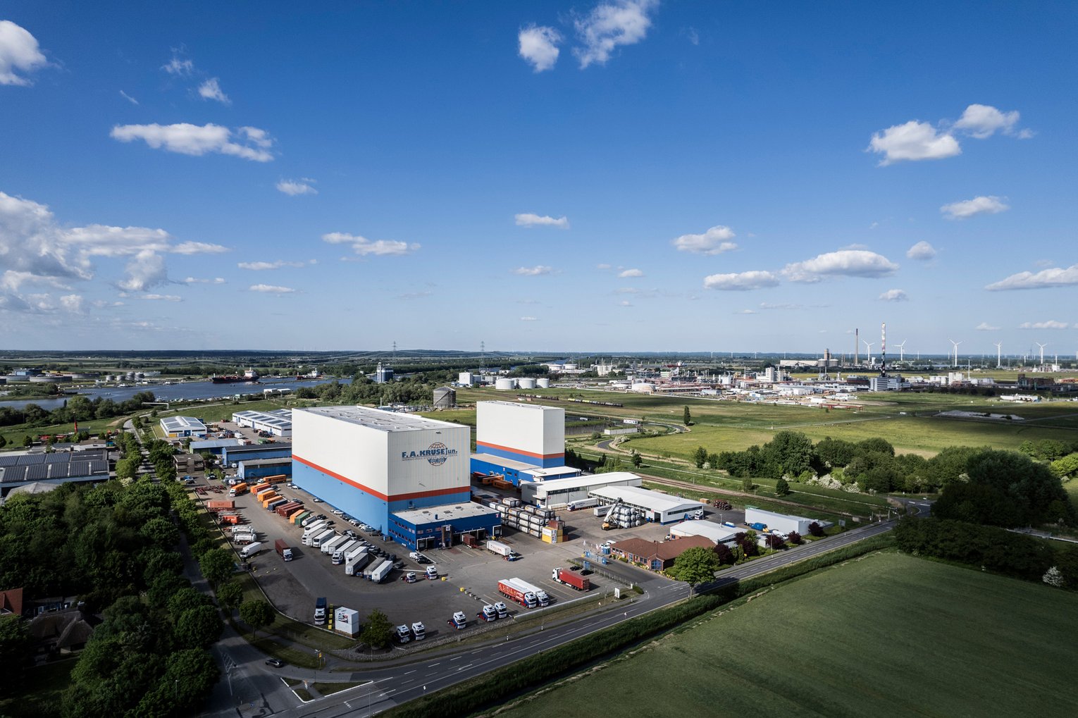 Luftaufnahme eines modernen Logistikzentrums mit zwei Hochregallagern, Parkplatz und Fluss im Hintergrund.